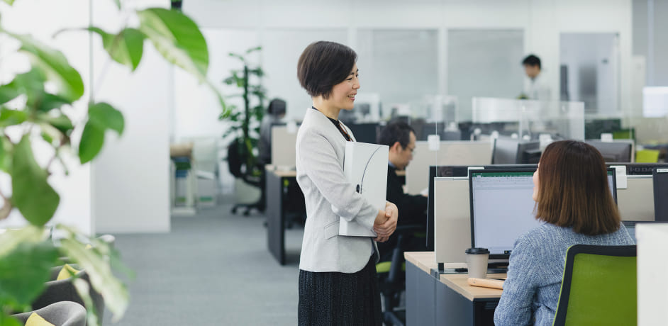 写真：フリーアドレス席に座っている社員に、別の社員が立ち寄って話しかけている様子